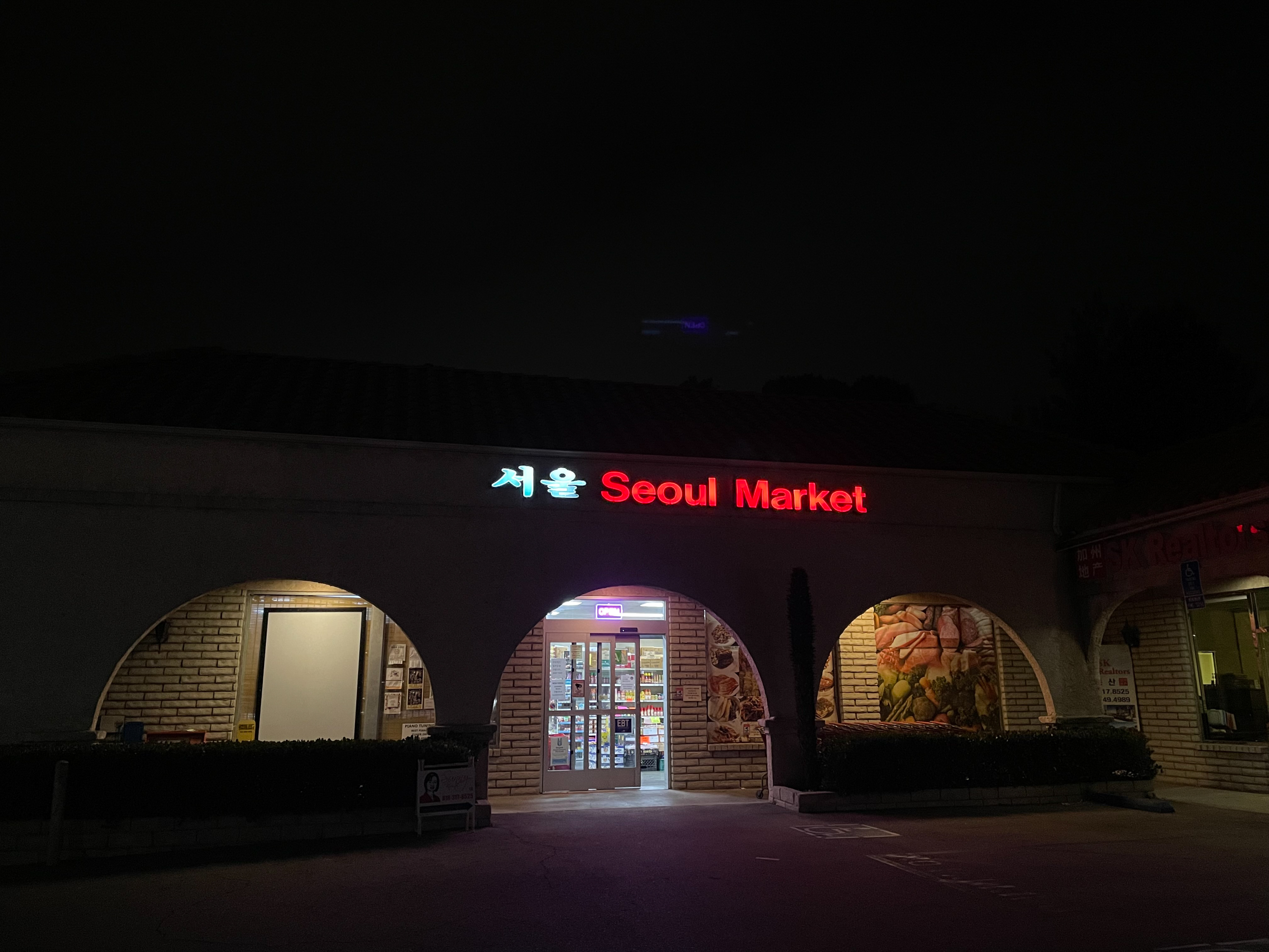 Exterior photo of a grocery store with neon letters on the front that reads Seoul Market. The sky is dark, the parking lot is empty, the rest of the strip mall is silent.